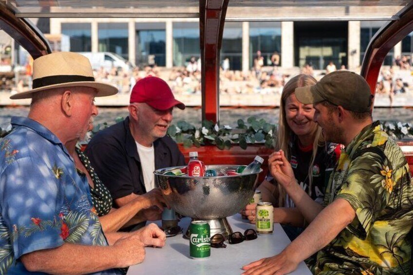 A group of people sitting at a table with a large metal bowl, drinks, and a cooler. 3