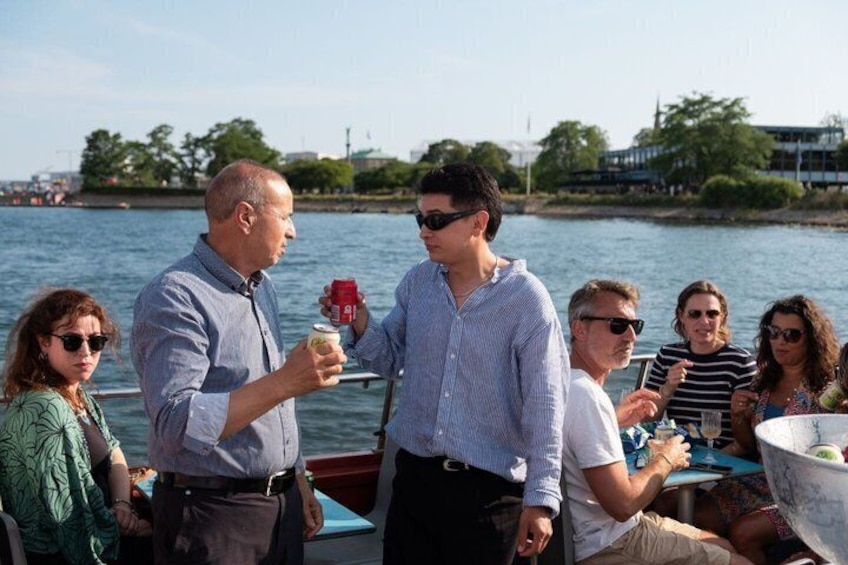 A group of people on a boat, some holding drinks, with a body of water and buildings in the background. 2