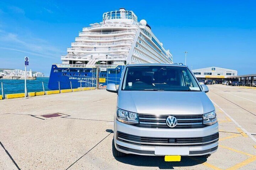 A silver Volkswagen van parked in a port area with a large cruise ship in the background. 4