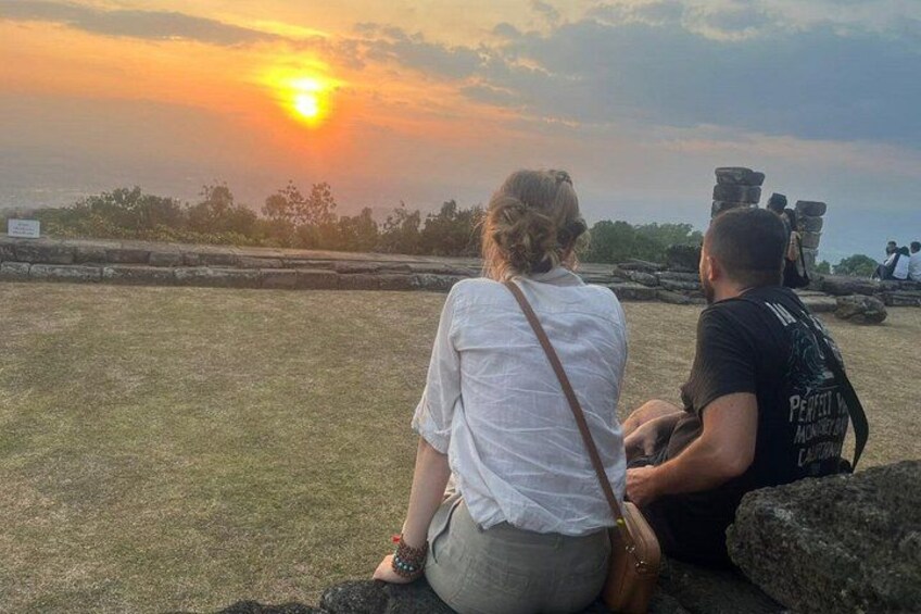Two people sitting on rocks, a sunset in the background, and a stone structure in the distance. 3