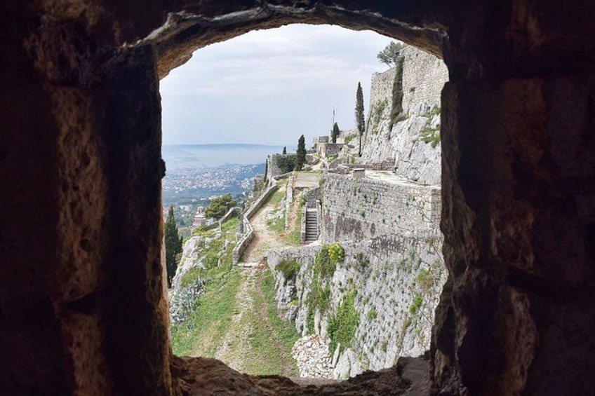 A historical stone structure with ruins, a pathway, and a distant view of a city and sea. 3