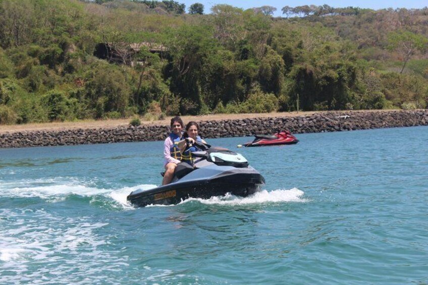 To personer på en jetski på vandet med en klippefyldt kystlinje og grøn bevoksning i baggrunden. 5