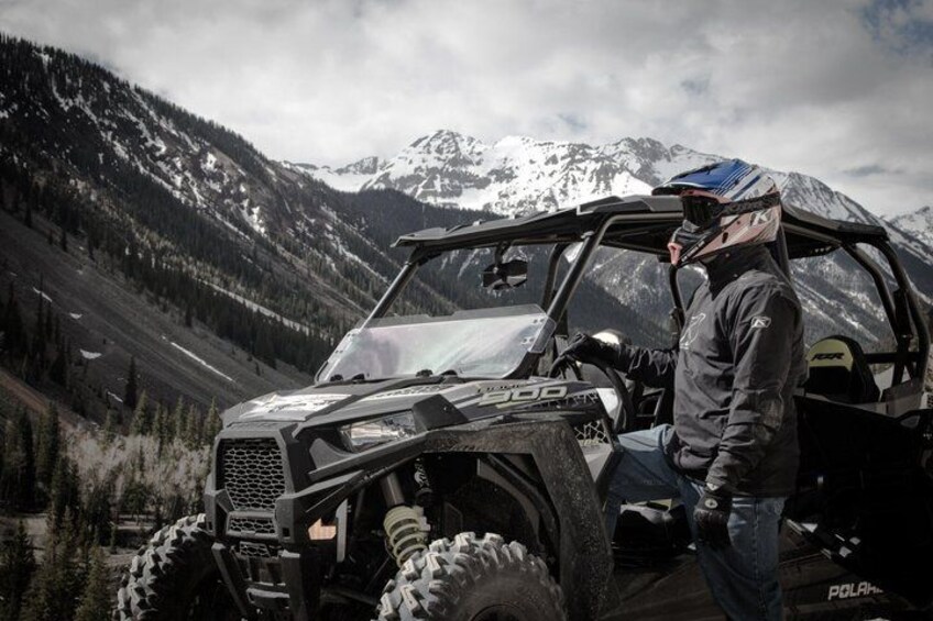 A person standing next to a black off-road vehicle with large tires, in a mountainous landscape. 2