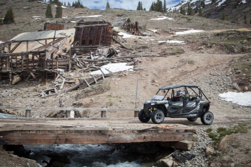 A vehicle crossing a makeshift wooden bridge over a river, with a mountainous landscape in the background. 4