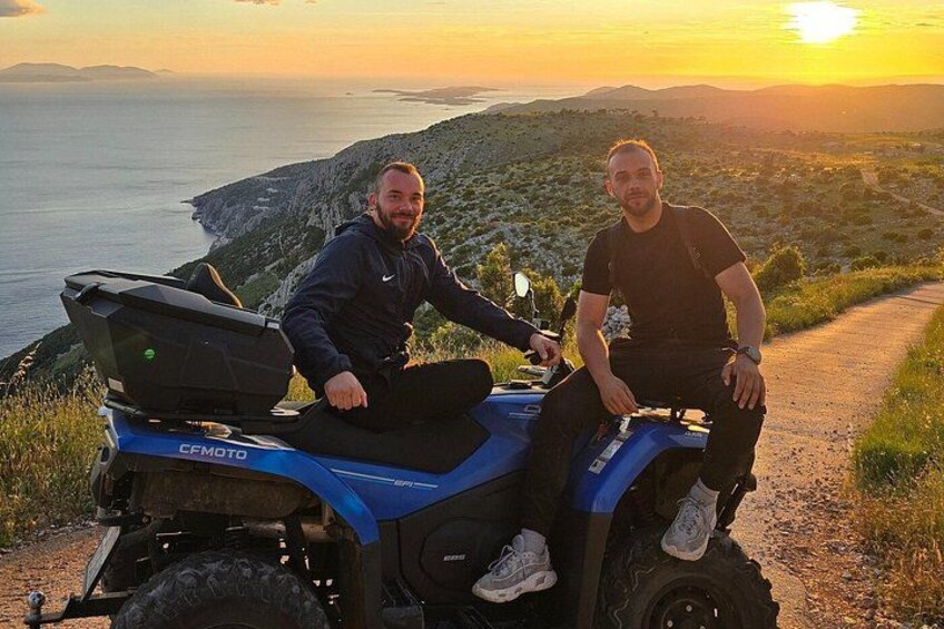 Two people on a blue ATV with a rugged landscape and a body of water in the background. 3