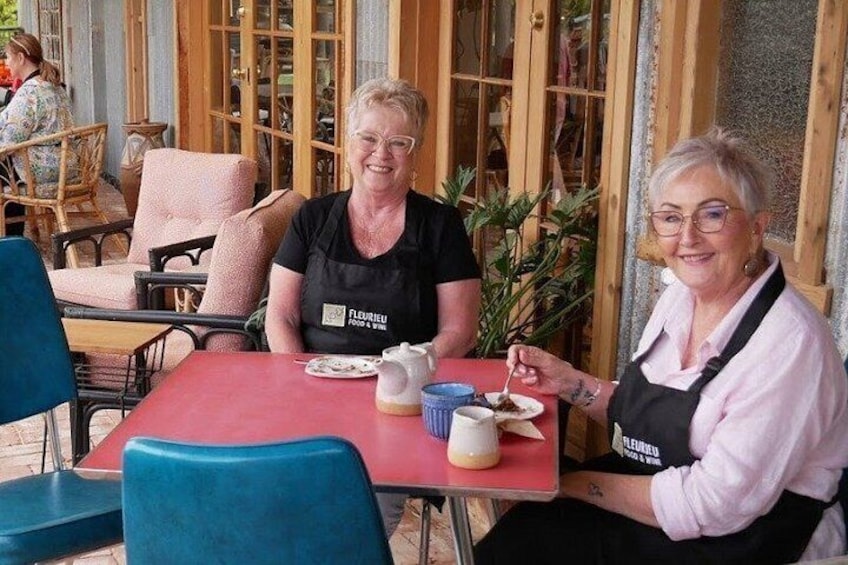 Two people sitting at a table with a red tablecloth, wearing aprons, and a plant in the background. 4