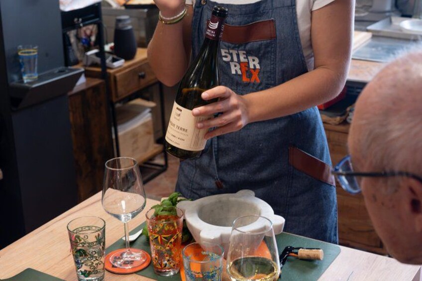 Una persona sosteniendo una botella de vino con etiqueta, de pie junto a una mesa con copas y un bol. 3