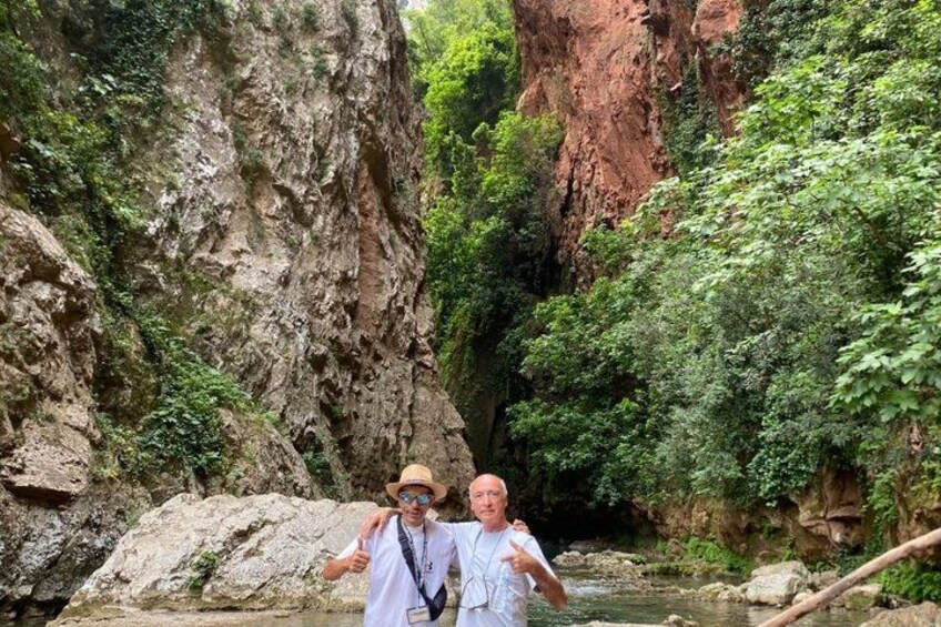 A rocky canyon with a river, greenery, and two people standing by the water. 4