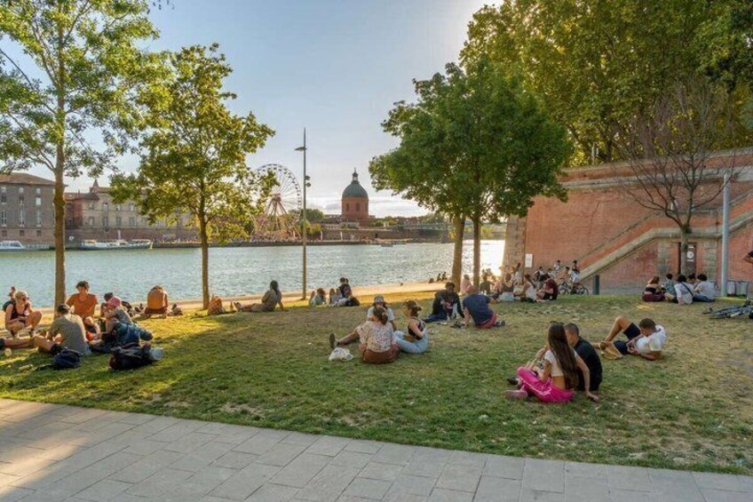 People sitting on grass by a waterfront with a Ferris wheel and buildings in the background. 3