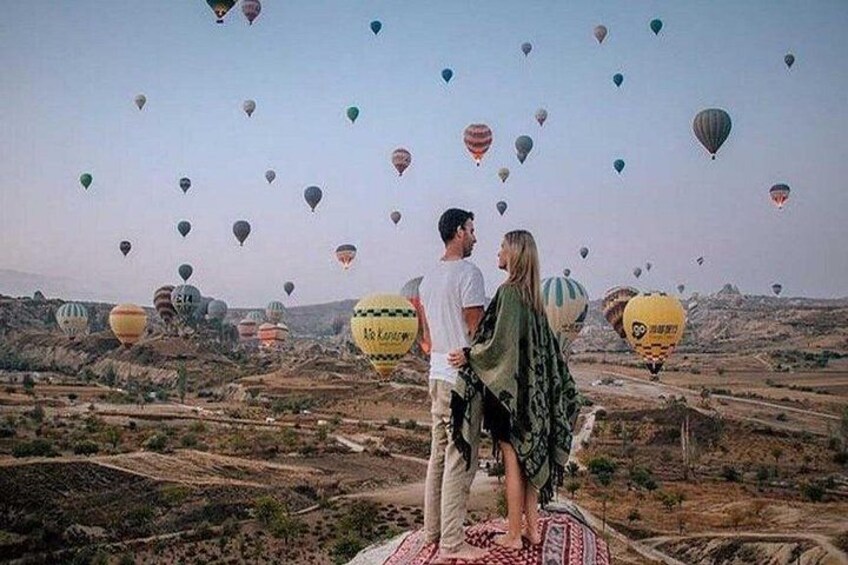 A couple standing on a blanket overlooking a landscape with hot air balloons. 1
