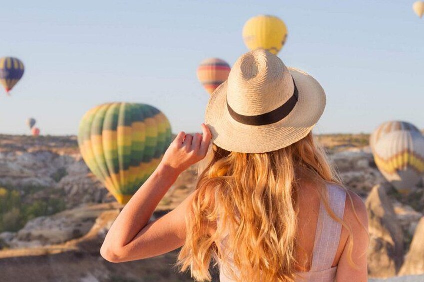 A person with long blonde hair wearing a straw hat, looking at colorful hot air balloons in the distance. 2