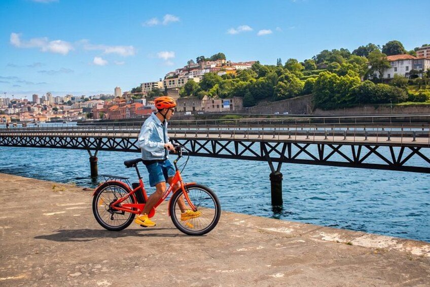 A person riding a red bicycle on a concrete path by a river, with a bridge and buildings in the background. 4