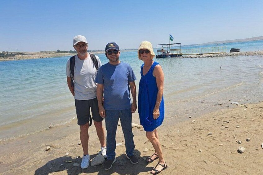 Three people standing on a sandy beach with a body of water and a pier in the background. 3