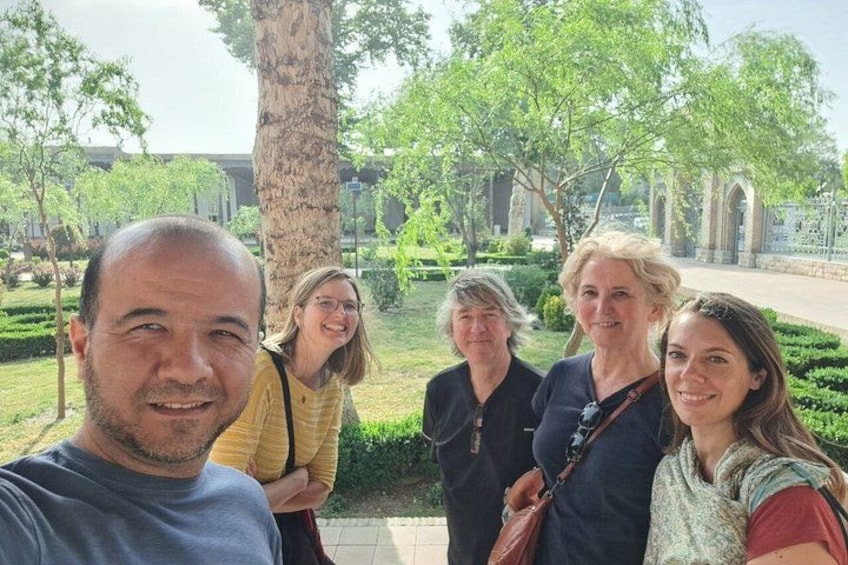 A group of people standing outdoors in a park with trees and a building in the background. 2