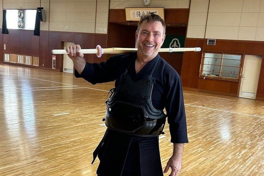 A person in a martial arts uniform holding a wooden staff in a gymnasium with wooden flooring and a mirrored wall. 4