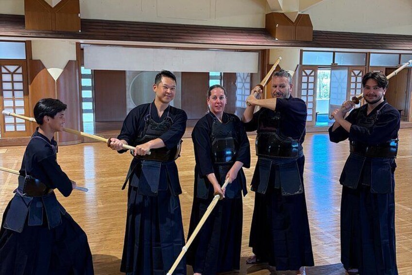 A group of people in traditional martial arts attire practicing with wooden staffs in a wooden-floored room with sliding doors. 4