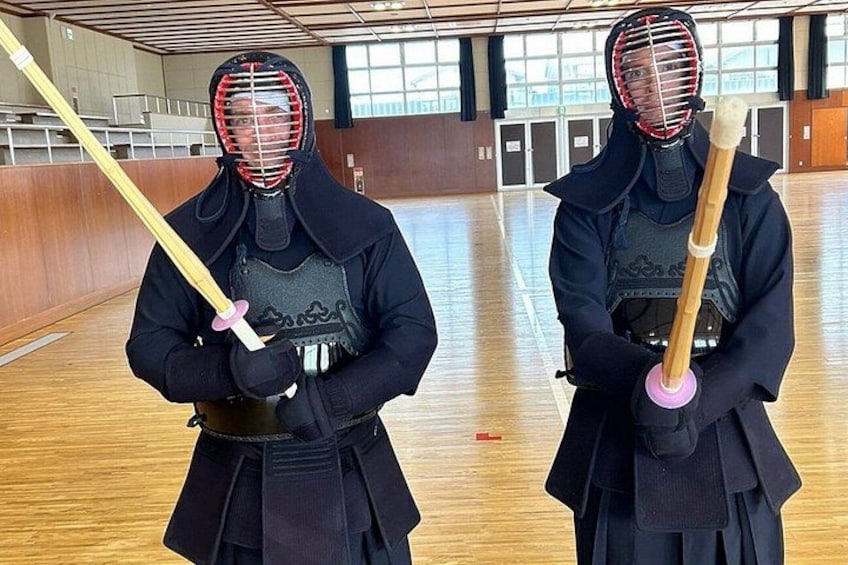 Two individuals in traditional martial arts attire holding wooden sticks, standing in a gymnasium with wooden floors and windows. 3