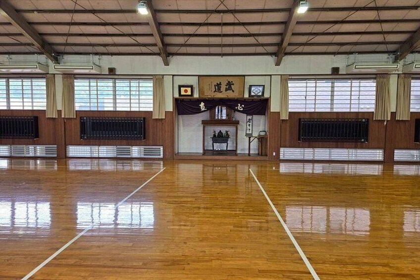 An indoor sports hall with a wooden floor, large windows, and a small stage with a banner. 3