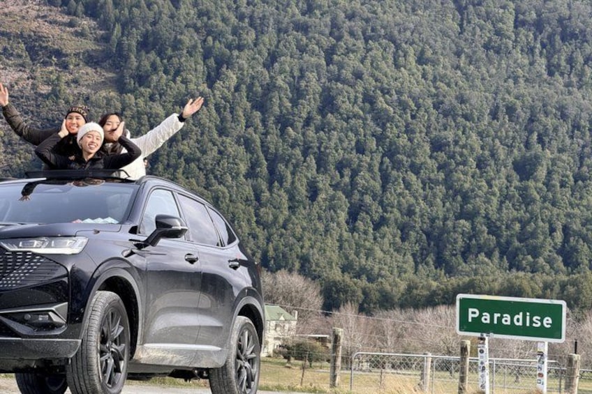 A car with people on top, a forested mountain in the background, and a road sign that reads 'Paradise'. 1