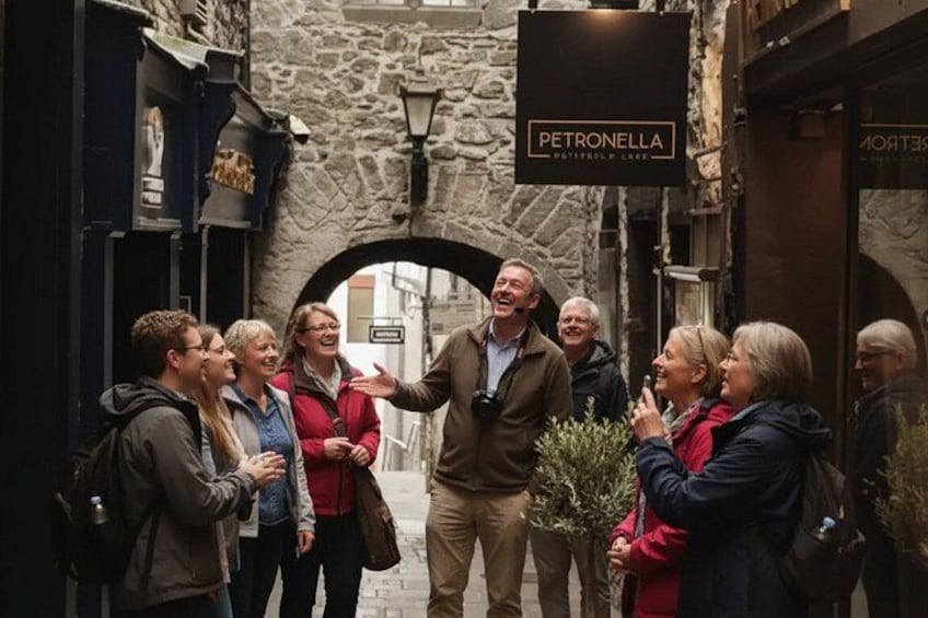 A group of people standing in a narrow street with stone walls and a sign for 'PETRONELLA' above a doorway. 5