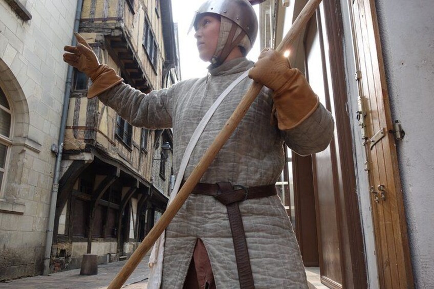 A person in medieval costume holding a long pole, standing in an urban alleyway with old buildings. 3