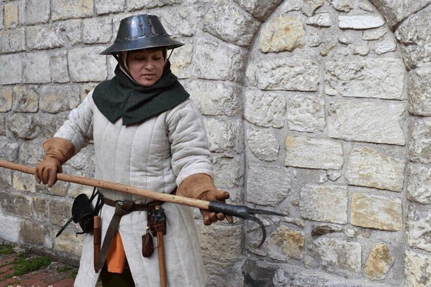 A person in medieval attire holding a long pole with a hook at the end, standing in front of a stone wall. 2