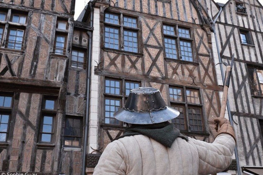 A person in historical costume with a hat and a cape, standing in front of a building with timber framing and multiple windows. 4
