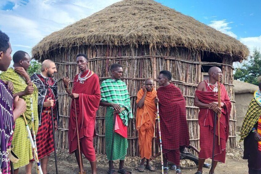 A group of people in traditional attire standing in front of a thatched hut. 1