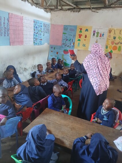A classroom with children seated at desks, a teacher holding a book, and educational posters on the walls. 3
