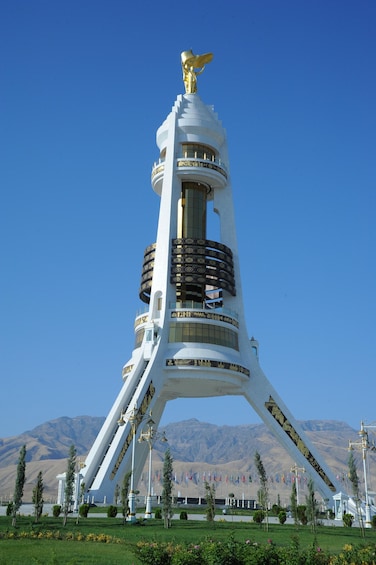 A tall white tower with a golden eagle statue on top, surrounded by flags and greenery. 5