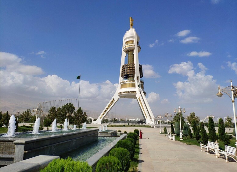 A tall white monument with a golden statue on top, surrounded by a fountain, trees, and a clear blue sky. 4