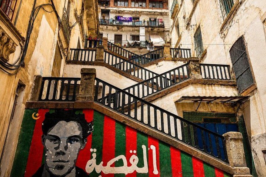 A narrow street with a staircase leading to an upper level, a mural with Arabic text, and a building with balconies. 4