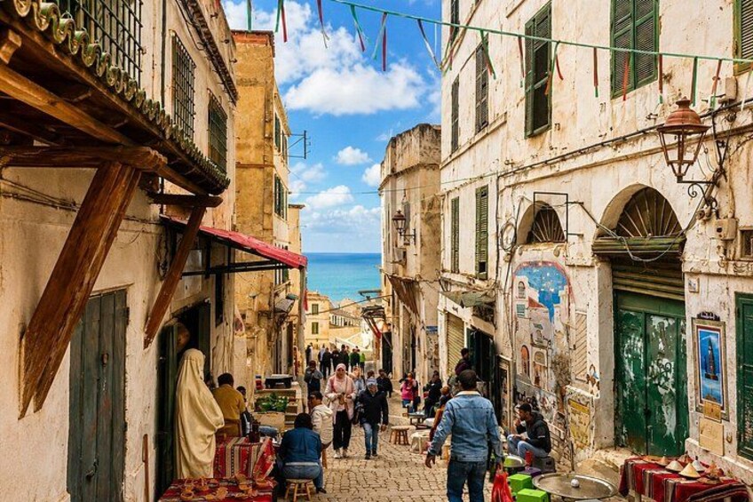 A narrow street with buildings, a blue sky, and a distant view of the sea. 3