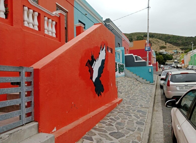 A colorful street with a red building featuring a black and white mural, parked cars, and a hilly landscape in the background. 2