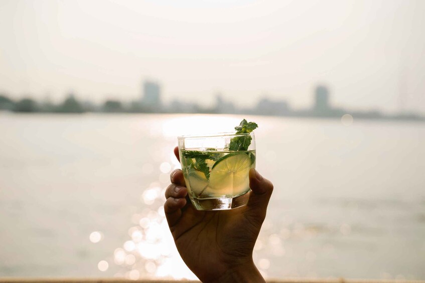 A hand holding a glass with a drink, a lime slice, and mint leaves, with a blurred city skyline and water in the background. 5