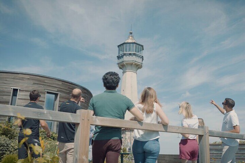 A group of people standing on a wooden deck overlooking a lighthouse and a curved metal structure. 3