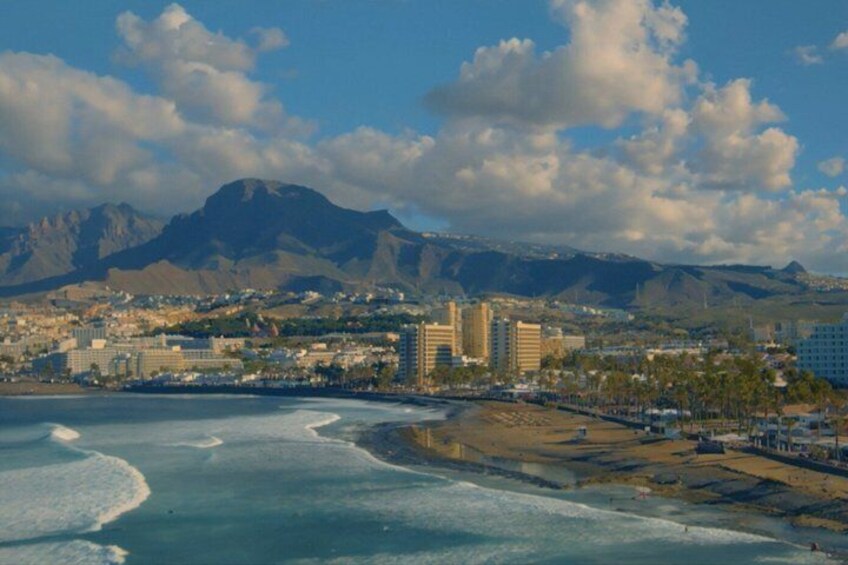 A coastal cityscape with mountains in the background, a beach, and several high-rise buildings. 1