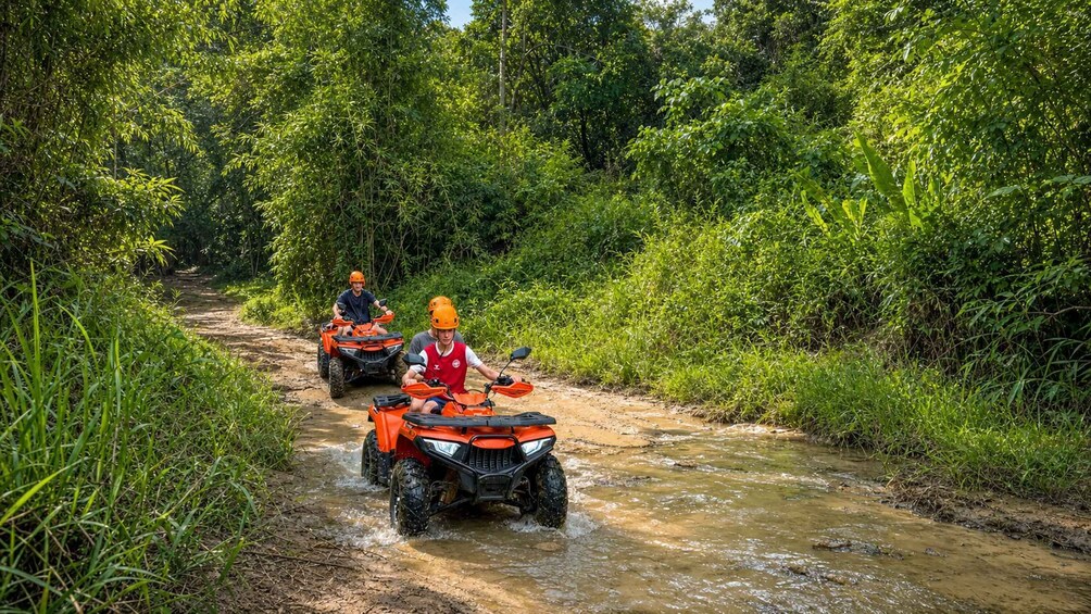 Two people on orange off-road vehicles navigating a muddy stream in a forested area. 4