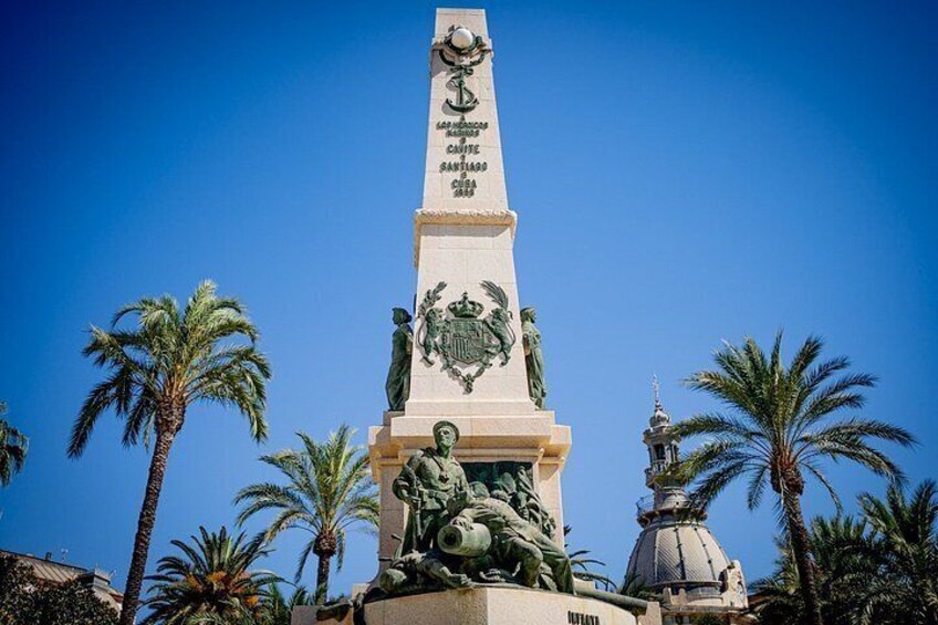 A tall monument with inscriptions, surrounded by palm trees and a dome structure. 2