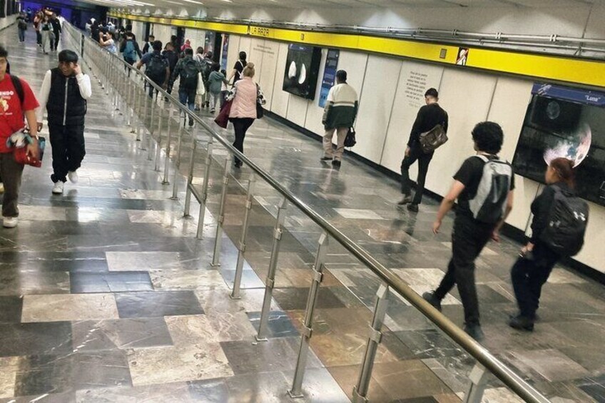 A subway station with tiled floors, metal railings, and people walking. 2