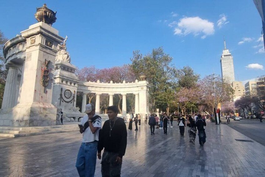 A public square with a monument, cherry blossoms, and a tall building in the background. 1