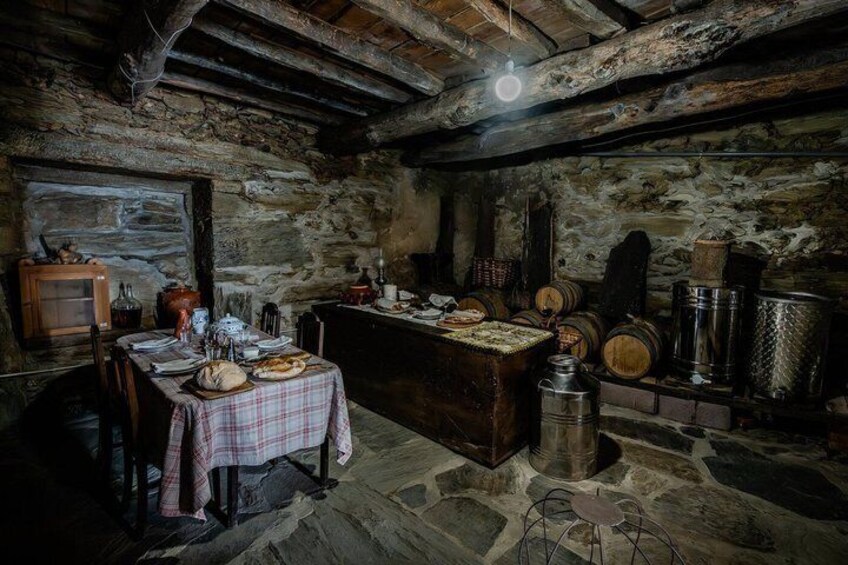A rustic stone kitchen with a wooden table set for a meal, surrounded by barrels and a large metal container. 3