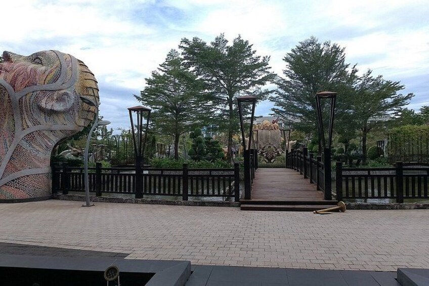 A paved pathway with a metal sculpture on the left, a wooden bridge in the center, and trees on either side. 3