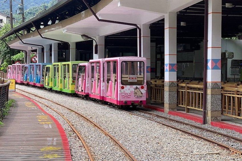 A colorful train with a pink front car, moving on tracks at a station with covered platforms and benches. 2