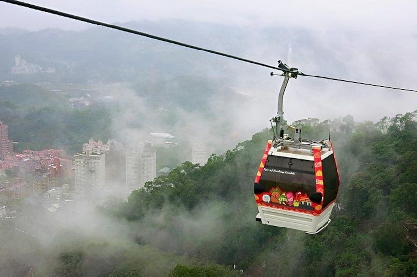 A cable car suspended over a forested area with buildings in the background. 2