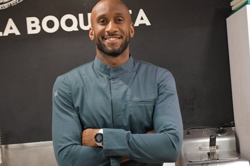 A person in a blue chef's jacket standing in front of a kitchen counter with a watch on their wrist. 1