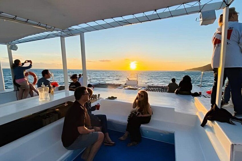 A boat with people sitting and standing, a sunset in the background, and a calm sea. 2