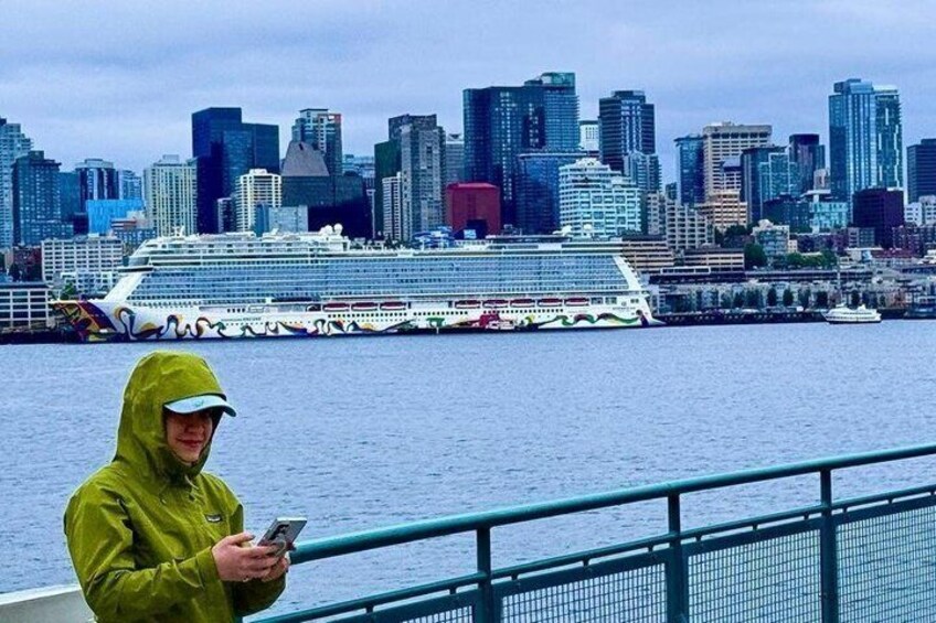 A person in a yellow raincoat standing on a boat with a city skyline and a cruise ship in the background. 1