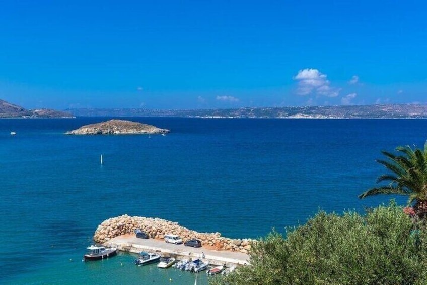 A coastal scene with a rocky island, clear blue sea, and a marina with boats. 1