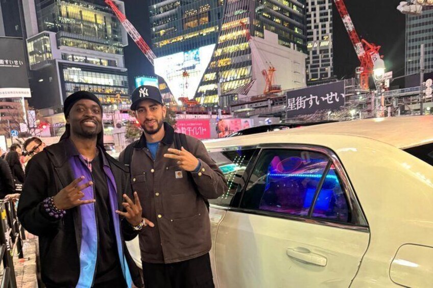 Two people standing in front of a white car with a blue interior, in a city with tall buildings and construction cranes. 3